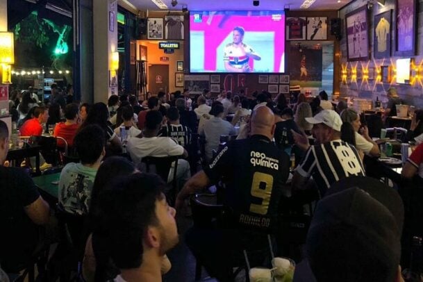 pessoas em um bar com telão assistindo um partida de futebol de São Paulo contra Corinthians